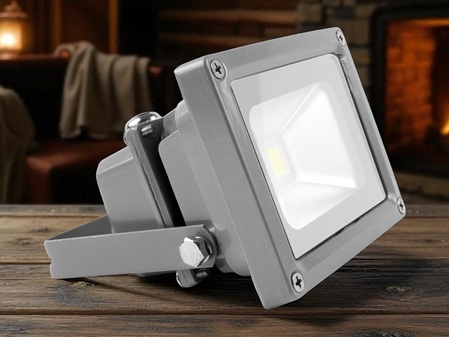 Grauer LED-Flutlichtstrahler mit kantigem Gehäuse und verstellbarem Haltebügel, auf einer Holzoberfläche platziert. In der Mitte der Leuchtfläche ist ein einzelner LED-Chip sichtbar. Im Hintergrund ein gemütlicher Innenraum mit Kamin und Sofa, in warmem Licht gehalten.