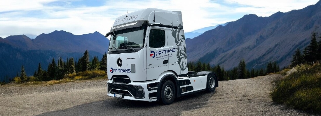 Ein weißer Sattelzug-Truck ohne Auflieger steht auf einer kurvigen Bergstraße mit Aussicht auf ein beeindruckendes Gebirgspanorama. Auf dem Truck befinden sich das Logo „PP-TRANS“ sowie eine stilisierte Wikingerzeichnung an der Seite. Über der Windschutzscheibe ist der Schriftzug „CharterWay“ zu lesen. Im Hintergrund ragen hohe, bewaldete Berge in den Himmel, und der Himmel ist leicht bewölkt mit einzelnen Sonnenstrahlen.