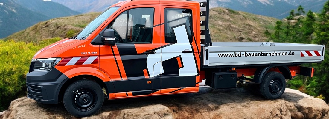 Ein rot-weißer Pritschenwagen mit grafischen Motiven und der Website www.bd-bauunternehmen.de parkt auf einem felsigen Gelände mit Bäumen und Bergen im Hintergrund.